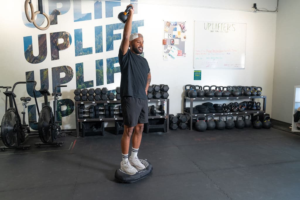 A trainer pressing a kettlebell overhead while standing on the board in a gym