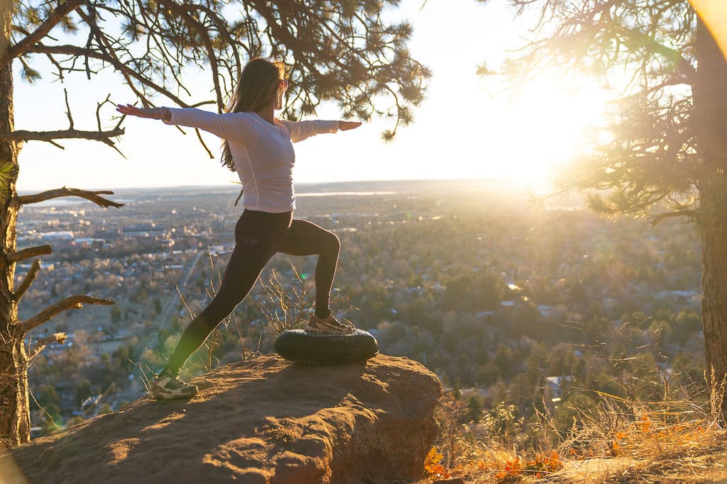 A woman holding warrior pose on a BALEROboard, on a rock above Boulder at golden hour